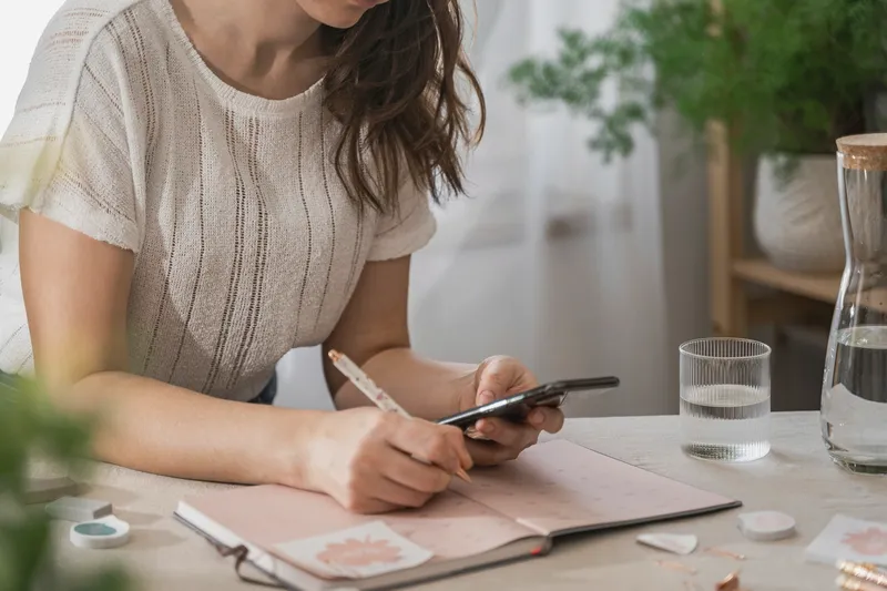 A woman at a table writing in a notebook, her phone in her other hand.